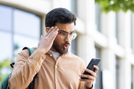 Upset student with phone in hand received notification with bad news, man with smartphone in hand outside university campus, nervous and disappointed.