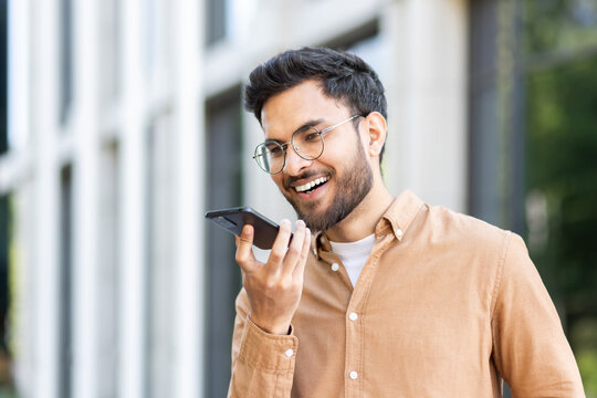A college student with glasses smiles as he uses the voice command function on his smartphone outdoors on campus