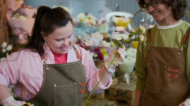 Tilt down view of young female intern with Down syndrome removing thorns from bush roses using thorn stripper with help of professional florist in flower shop