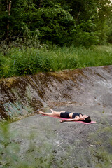 Yoga trainer lying on pink mat, one leg extended straight up, other leg extended horizontally, deep stretch and flexibility in lush green outdoor setting