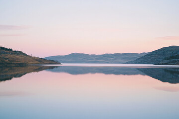 Fototapeta premium A tranquil mountain lake at sunrise with calm, glass-like reflections and soft pastel sky hues.