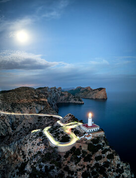 Aerial image of light house on dramatic cliff coastline with curvy road illuminated by car lights during blue hour, at Far the Formentor, Mallorca, Spain 