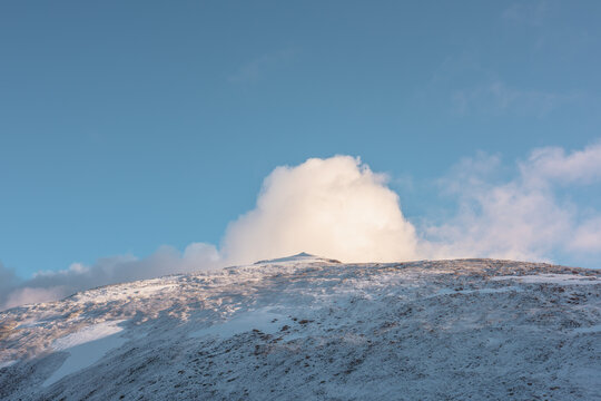 Lush sunlit low cloud on snow-capped mountain top at sunrise. Snowy stone hill in sunlight at early morning. High mountain in freshly fallen snow. Snow-covered stony peak and fluffy cloud in blue sky.