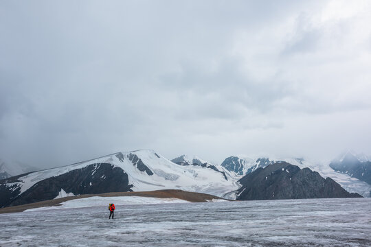 Man on ice pass with view to big glacier tongue among sharp rocks and large snow-capped mountains in rainy low clouds. Guy against dark atmospheric mountain silhouettes in rain in gray cloudy sky.
