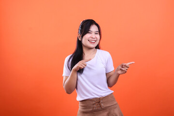 Fototapeta premium A woman looks at the camera wearing a white shirt and brown skirt with both hands pointing to the right while smiling, isolated on an orange background