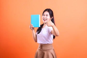 Naklejka premium A woman with black hair holding a blue book and giving thumbs up against orange background