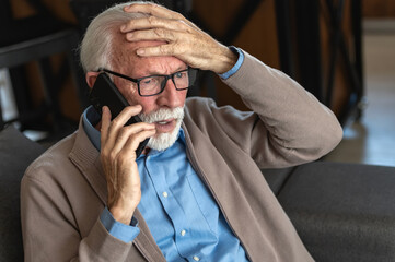 An elderly man with glasses and a beard, wearing a blue shirt and beige cardigan, looks distressed...