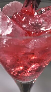 fresh red drink pouring into a cocktail glass with ice cubes on a gray sunny restaurant tabletop, slow motion