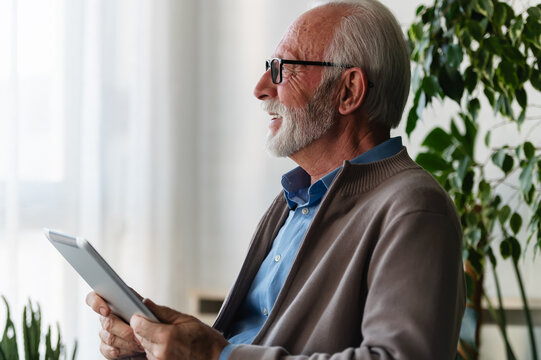 An elderly man with glasses and a beard sits by a window, holding a tablet and looking outside thoughtfully, surrounded by plants 