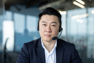 Portrait of an Asian young man wearing a headset and a suit sitting in the office at the workplace...