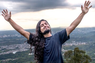 Man with long hair and a beard raising his arms in joy while standing on a mountain top with a...