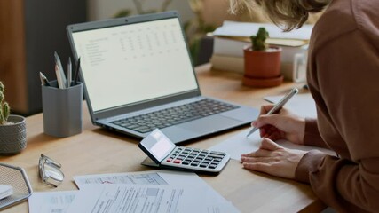 Medium shot of hands and midsection of anonymous young woman sitting at desk at home, looking through invoices, adding up monthly utility payments on calculator, while managing expenses
