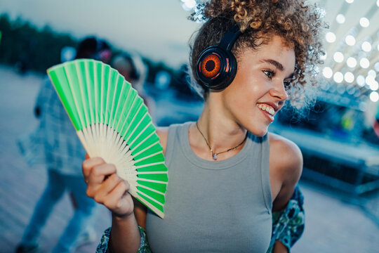Young curly woman enjoying music at summer open air festival using headphones and waving green fan
