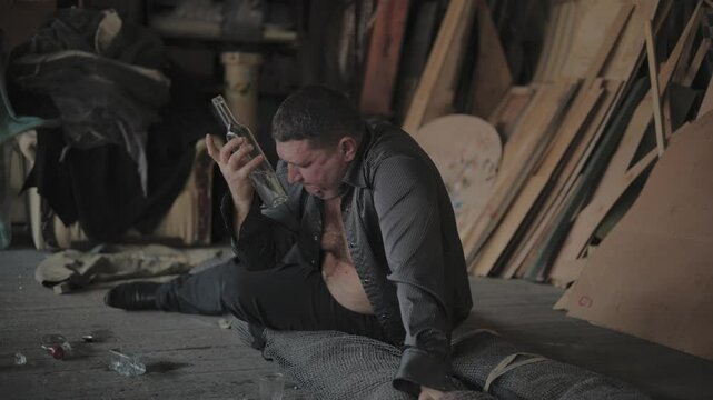 The concept of a man addict in a deep crisis.Drunk man in a gray T-shirt sitting on the floor.Alcoholic problems, addiction.The guy is drinking vodka from a bottle.
