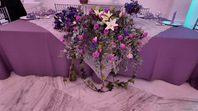 A table is covered with a floral arrangement of purple and white flowers. The table is set for a special occasion, and the flowers are the centerpiece