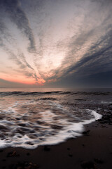 A wave crashing in a circular motion on a dark sand beach underneath a sunrise sky. This photo is looking over Buzzards Bay on a winter morning at dawn, off the coast of Massachusetts.