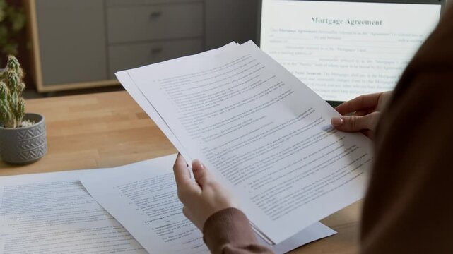 Over-shoulder shot of hands of unrecognizable female sitting at desk at home, leafing through pages of paper copy of mortgage agreement and reading contract text, while planning to buy new house