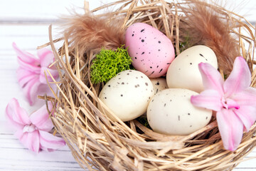 Easter decor on the white wooden table - nest with speckled pink and beige eggs, moss and feathers and sprinkled pink hyacinth flowers around.