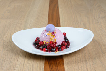 A scoop of purple ice cream with assorted berries is arranged on a white plate on a wood background