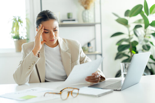Frustrated caucasian business woman, looking stressed because of results of work, holding her head while reading a document at her desk, feeling confused, upset and disappointment