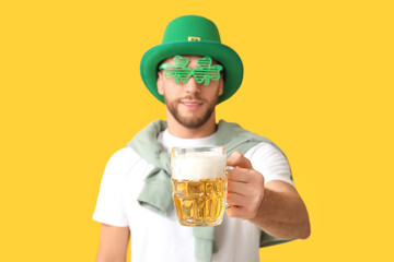 Young man in leprechaun hat and decorative glasses in shape of clover with glass of beer on yellow...