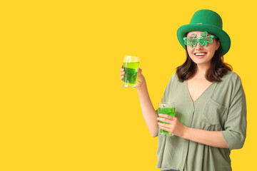 Beautiful young woman in leprechaun hat with glasses of beer on yellow background. St. Patrick's...