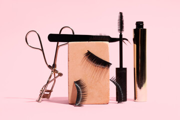Plaster podium with false eyelashes, curler, tweezers and mascara on pink background