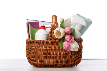 Wicker basket with bottles of detergents, cleaning tools and tulip flowers on table against white...