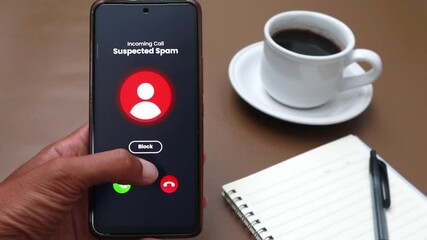 Hand holding smartphone receiving suspected spam call. On the brown table there is a cup of coffee and a notebook - Powered by Adobe