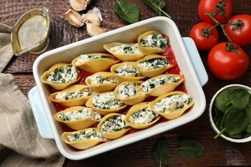 Delicious conchiglie pasta with ricotta cheese, spinach and tomato sauce in baking dish on wooden table, flat lay