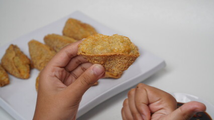 One hand is holding fried tahu walik aci, there is a plate of tofu walik with soy sauce and chili sauce beside it, placed on a white base and background (isolated white).