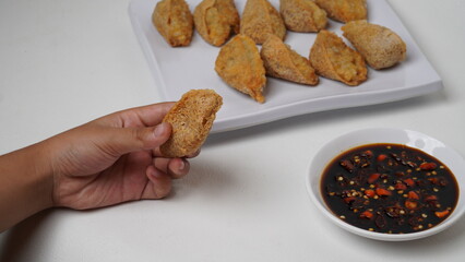 One hand is holding fried tahu walik aci, there is a plate of tofu walik with soy sauce and chili sauce beside it, placed on a white base and background (isolated white).