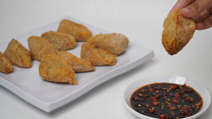 One hand is holding fried tahu walik aci, there is a plate of tofu walik with soy sauce and chili sauce beside it, placed on a white base and background (isolated white).