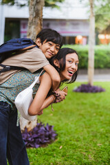 Fototapeta premium Portrait of Asian teenage sister carrying her little brother on her back in a park outdoors with happy expression on their faces