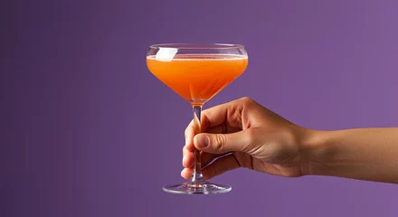 Cocktail in glass held in hand on purple background. Cocktail drink in coupe glass
