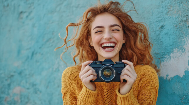 cheerful woman with curly hair joyfully laughs while holding camera against blue wall. Her vibrant yellow sweater adds pop of color to scene, capturing moment of happiness