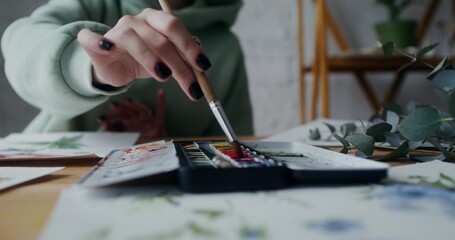 A female artist mixes watercolors with a brush to get the right color while painting wild flowers on paper. Close-up of female hands with brush