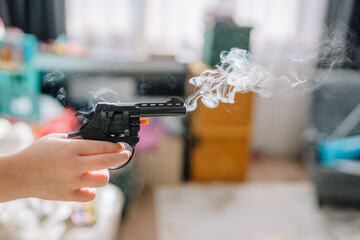 A child’s hand holding a toy revolver with smoke coming from the barrel in a blurred indoor...
