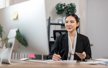 Young committed asian business the data analyst woman counts money in hands sitting  with a desktop computer in the office.
