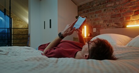A young man in glasses dressed in casual clothes is typing text on a mobile phone, while lying across the bed in the evening in a stylish home interior