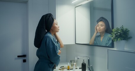 Naklejka premium Young beautiful Asian woman in a bathrobe, with a towel on her head, touches her face while looking at herself in the mirror in a bright stylish bathroom
