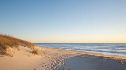 Peaceful sandy beach with ocean waves and dunes covered in grass at sunrise. The sky displays soft hues, creating a tranquil atmosphere