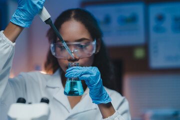 Young female scientist wearing protective glasses and gloves is carefully adding reagent to...