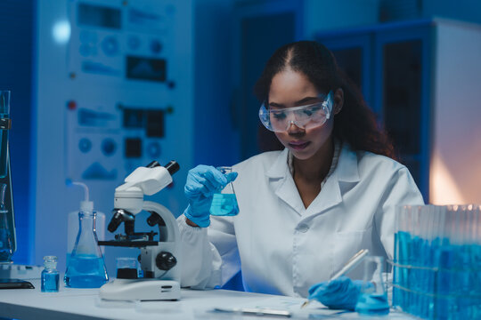 Young female scientist wearing lab coat and safety goggles holding beaker with blue liquid reagent, writing notes and using microscope in modern laboratory