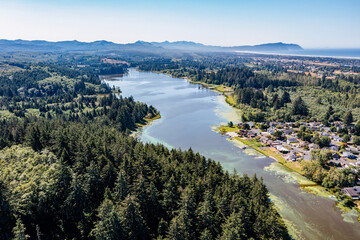 Aerial drone view of Cullaby Lake near Warrenton, Oregon, featuring a lakeside residential area surrounded by lush forests, with the Pacific Ocean, scenic wetlands, and Highway 101 in the background