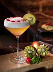 A beautifully crafted yellow cocktail in a wide coupe glass, topped with a creamy foam layer and decorated with red syrup drops.