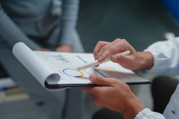 Businessman in lab coat holding clipboard and pointing with pen at charts while engaging in discussion about financial results with colleague, emphasizing strategy and growth