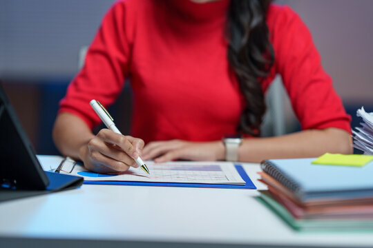Focused businesswoman in a red blouse writing on financial documents at a desk, surrounded by office supplies and a digital tablet, emphasizing productivity and organization - Powered by Adobe