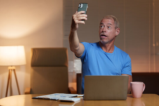Surprised mature man taking a selfie with his smartphone while working late at night in his home office, using a laptop and reviewing documents