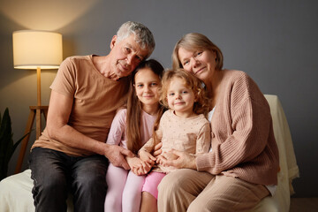 A loving family sits closely together on a couch, with a grandfather, grandmother, and two young...